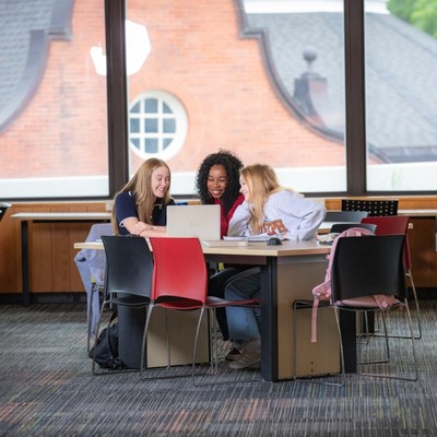 Three students sit at a table in the McLaughlin Library looking at a laptop together smiling.