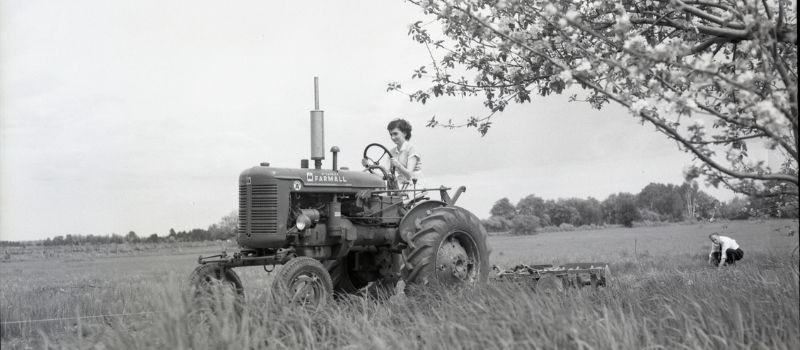 A woman farmer riding a tractor across a field with a man bent down watching her from afar.
