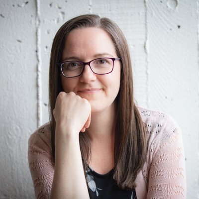 Amber Allen sits in front of a white concrete wall, wearing glasses, and rests her chin on her hand, smiling slightly to the camera.