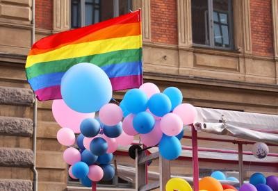 Balloons and flags fly at Pride celebration. The balloons are trans-colours, pink and blue. The flag is the Pride flag.