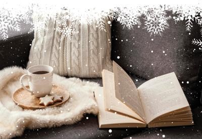 An open book sits on a couch  next to a cup of coffee and some snowflake-shaped cookies sitting on a fuzzy blanket.